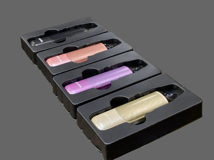 Four vape devices in black retail packaging, shown in different colors: black, coral pink, purple, and beige, arranged diagonally inside a glass display case.
